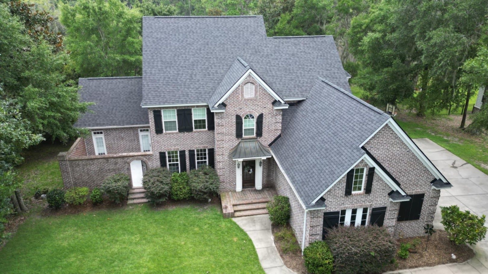 Two-story brick house with dark gray roof, black shutters, and green lawn, trees in background.