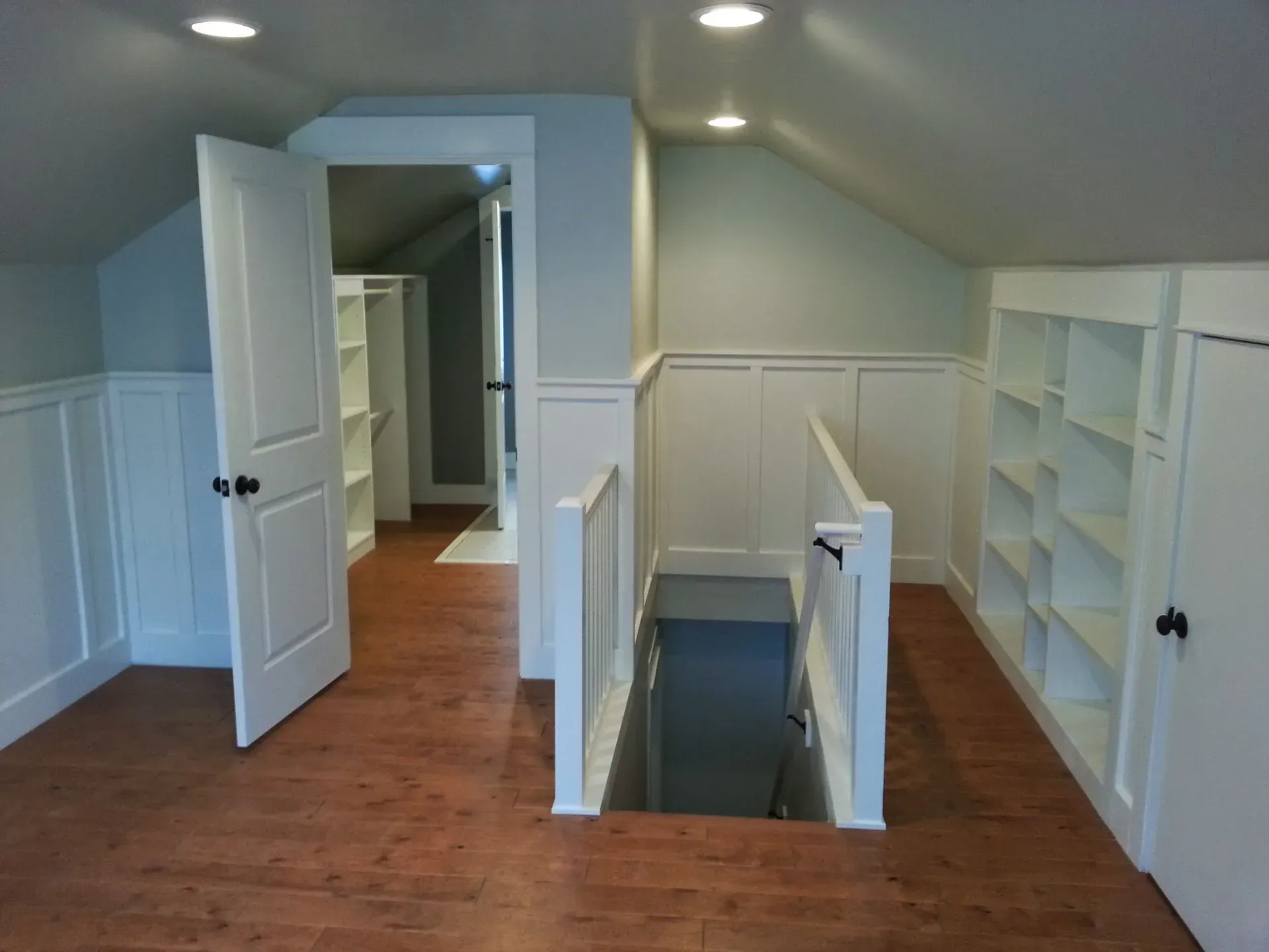 Finished attic room with white shelves, wood floor, and an open doorway by a stair opening.