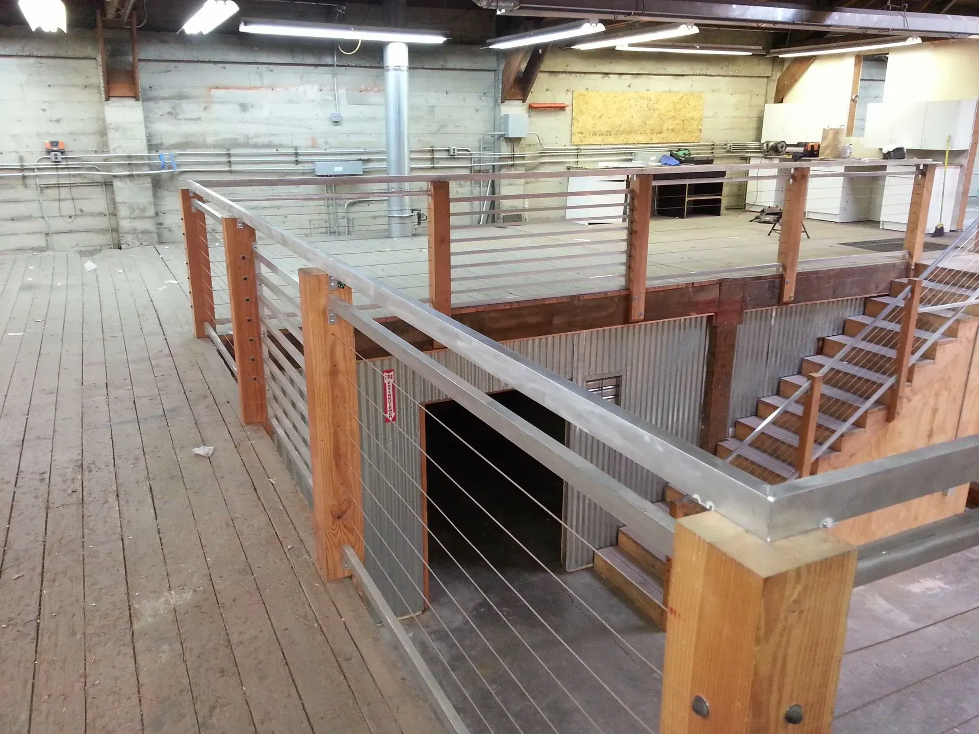 Interior of a warehouse mezzanine with metal railing, stairs, and wooden posts around an open lower level