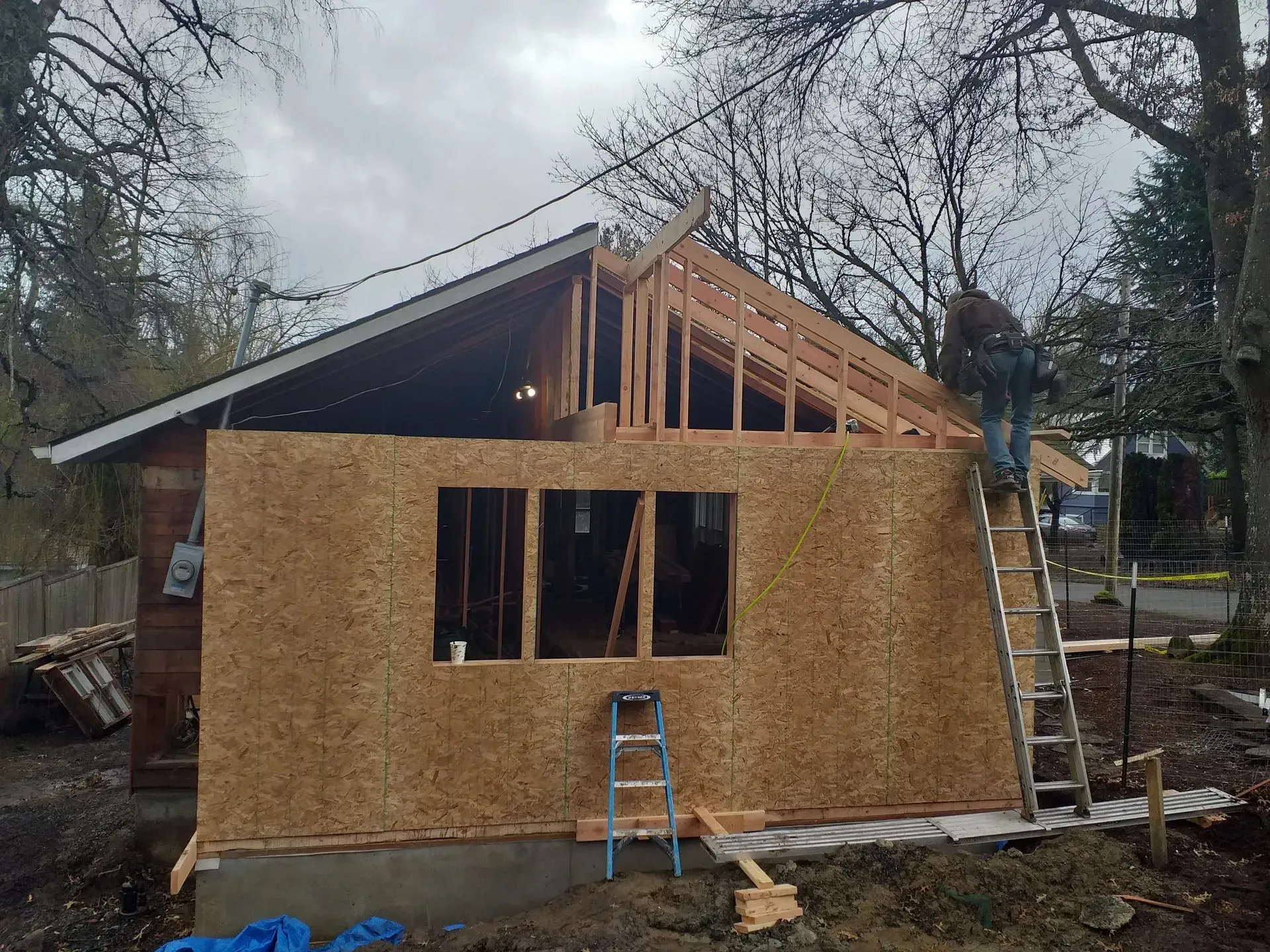 Wood-framed house under construction with plywood walls, exposed rafters, and a worker on a ladder.