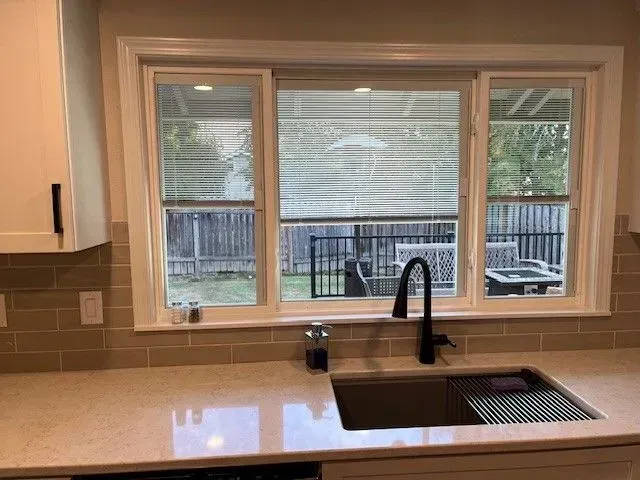 Kitchen with white cabinets, beige countertops, black faucet, and window above a sink overlooking a backyard.