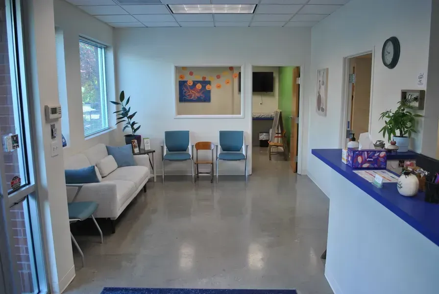 Bright waiting room with blue chairs, white walls, a reception counter, and potted plants.