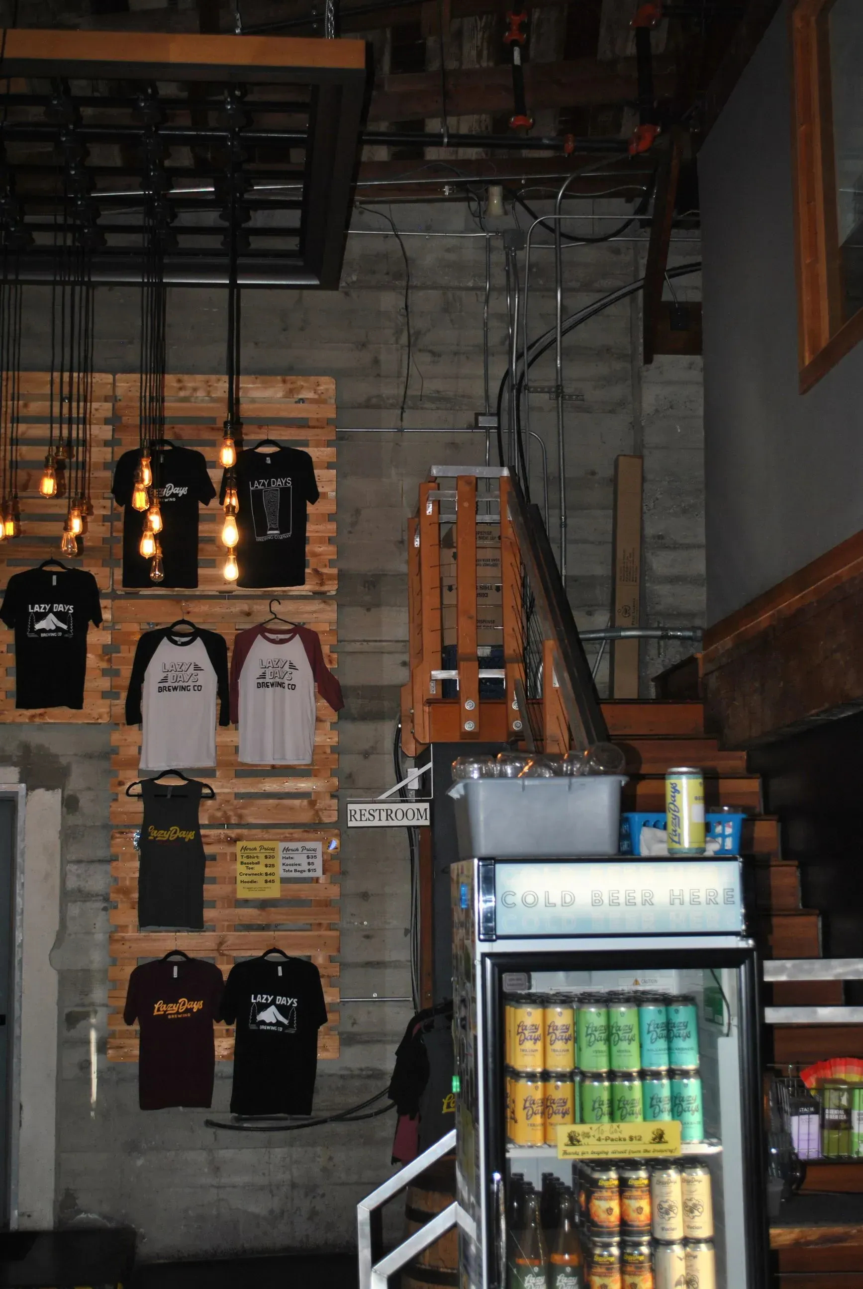 Interior of a shop with hanging T-shirts, a staircase, and a refrigerated drink display.