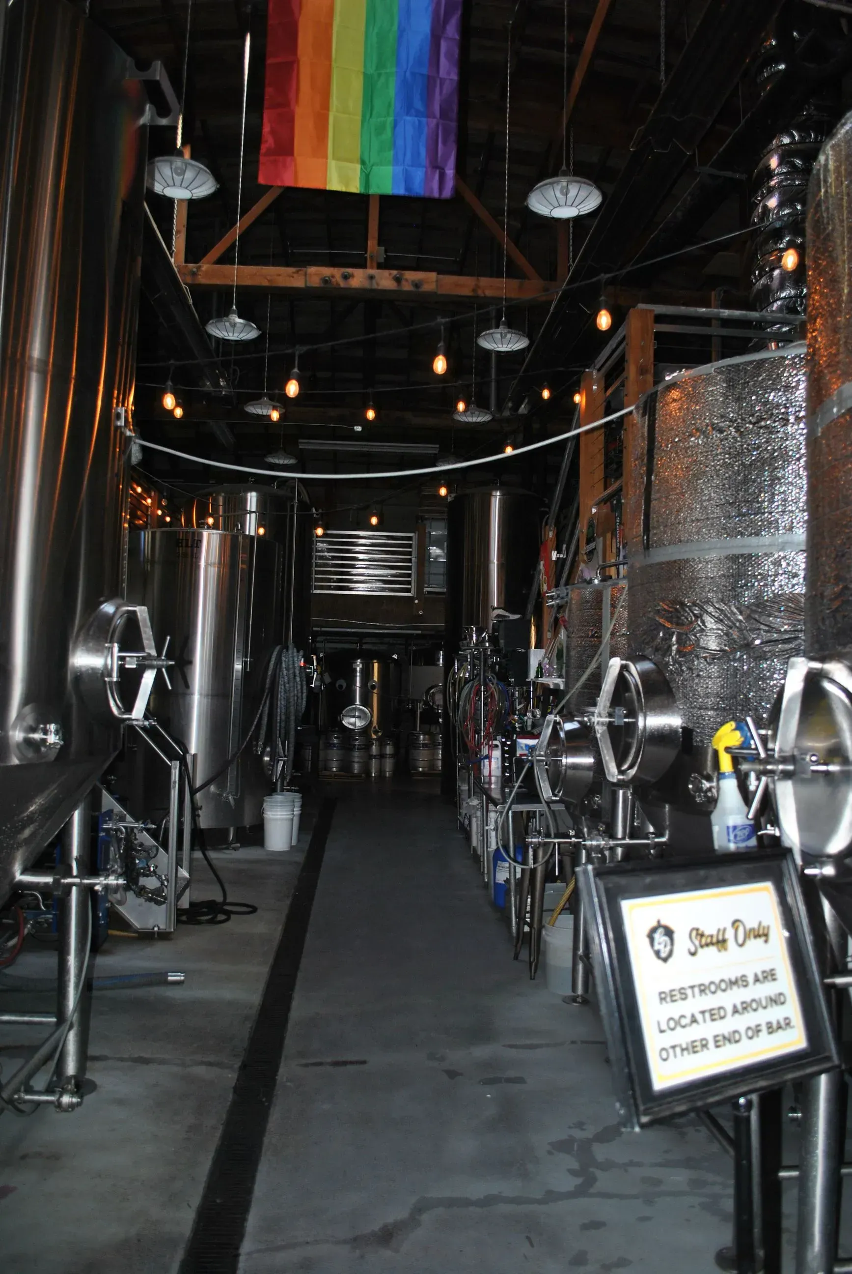 Industrial brewery interior with stainless tanks, taps, and a rainbow Pride flag overhead