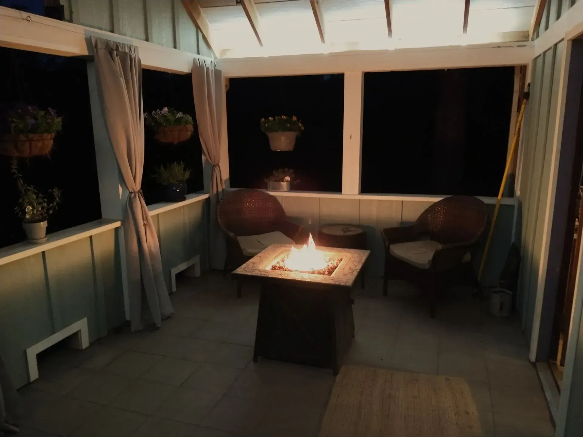 Cozy screened porch with wicker chairs, a central fire table, and potted plants at night