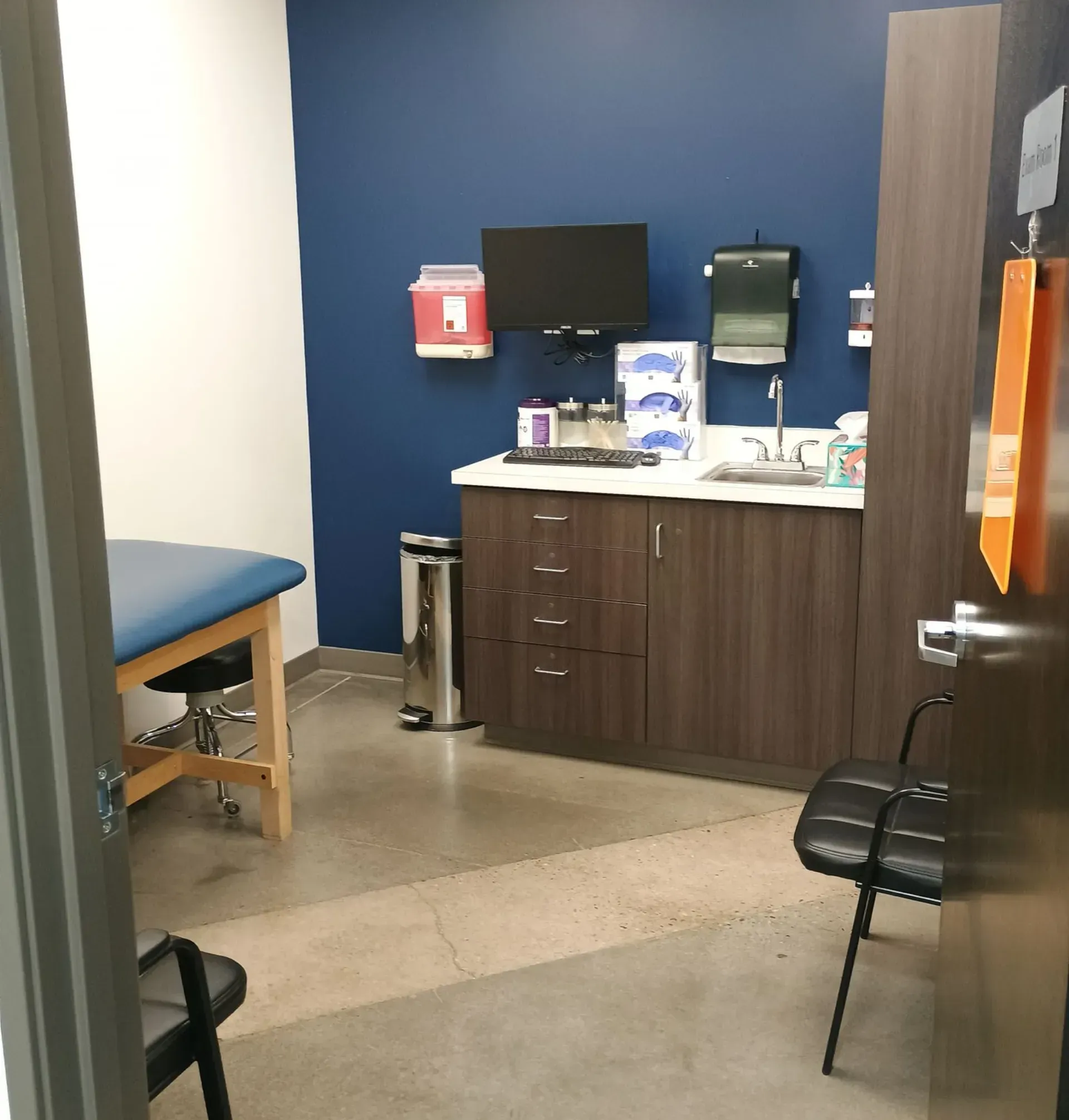 Clinic exam room with blue walls, exam table, sink, cabinets, and medical supplies