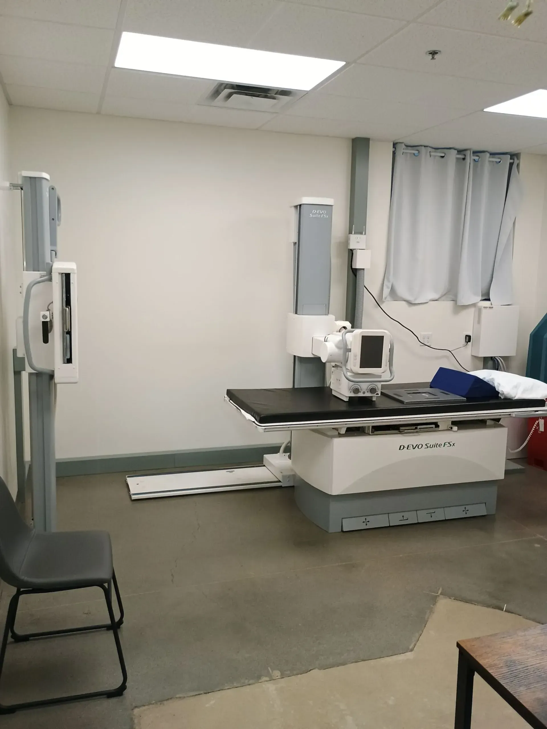 Empty medical exam room with X-ray table, chair, and curtains under bright fluorescent lights