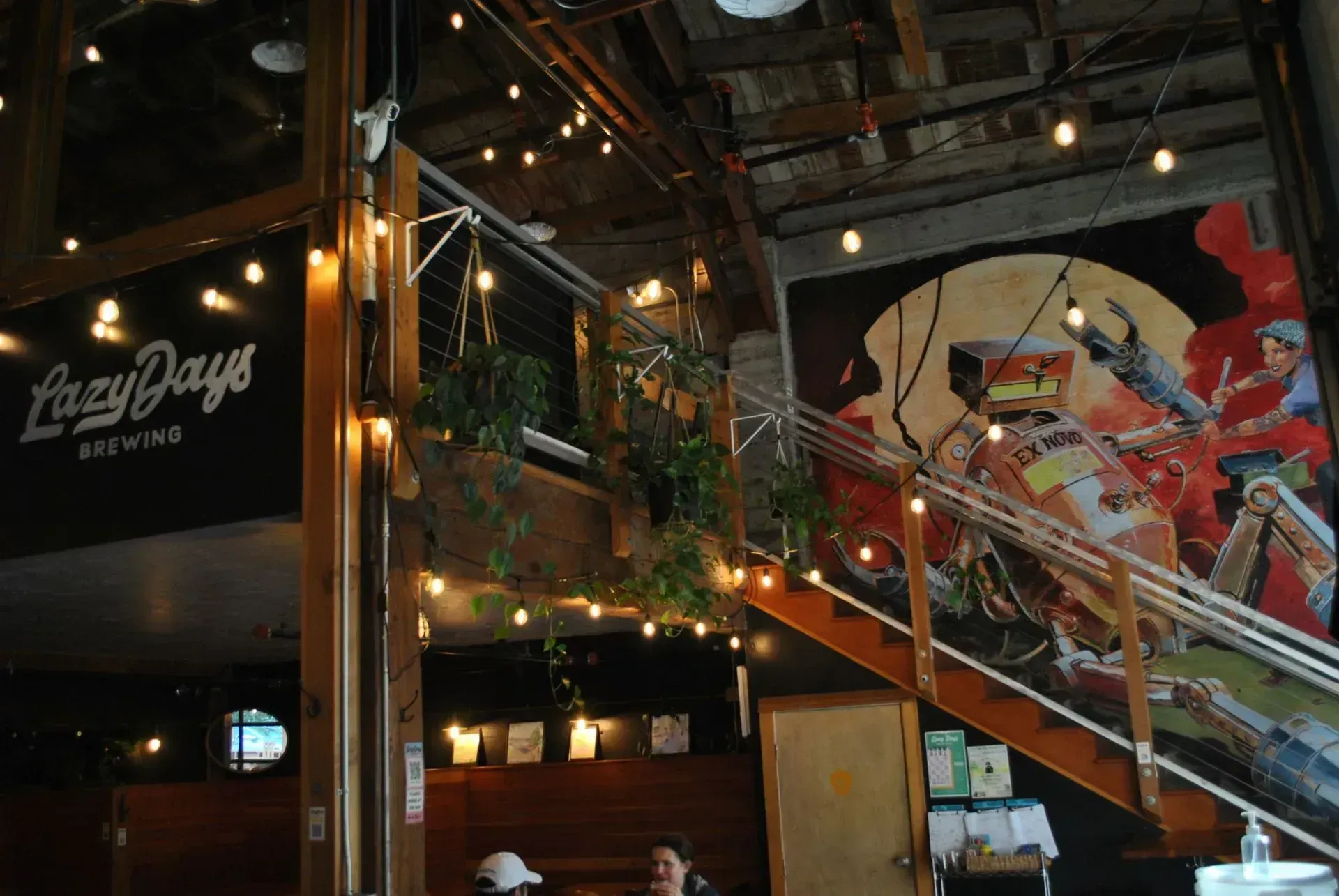 Indoor brewery with string lights, wooden stairs, and a large colorful mural on the wall
