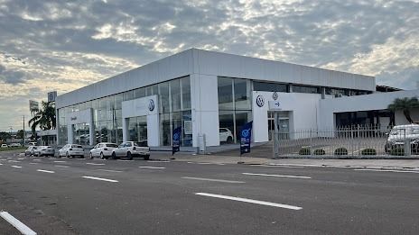 Exterior de uma concessionária de veículos, prédio branco, carros em exposição, céu nublado, rua em frente.