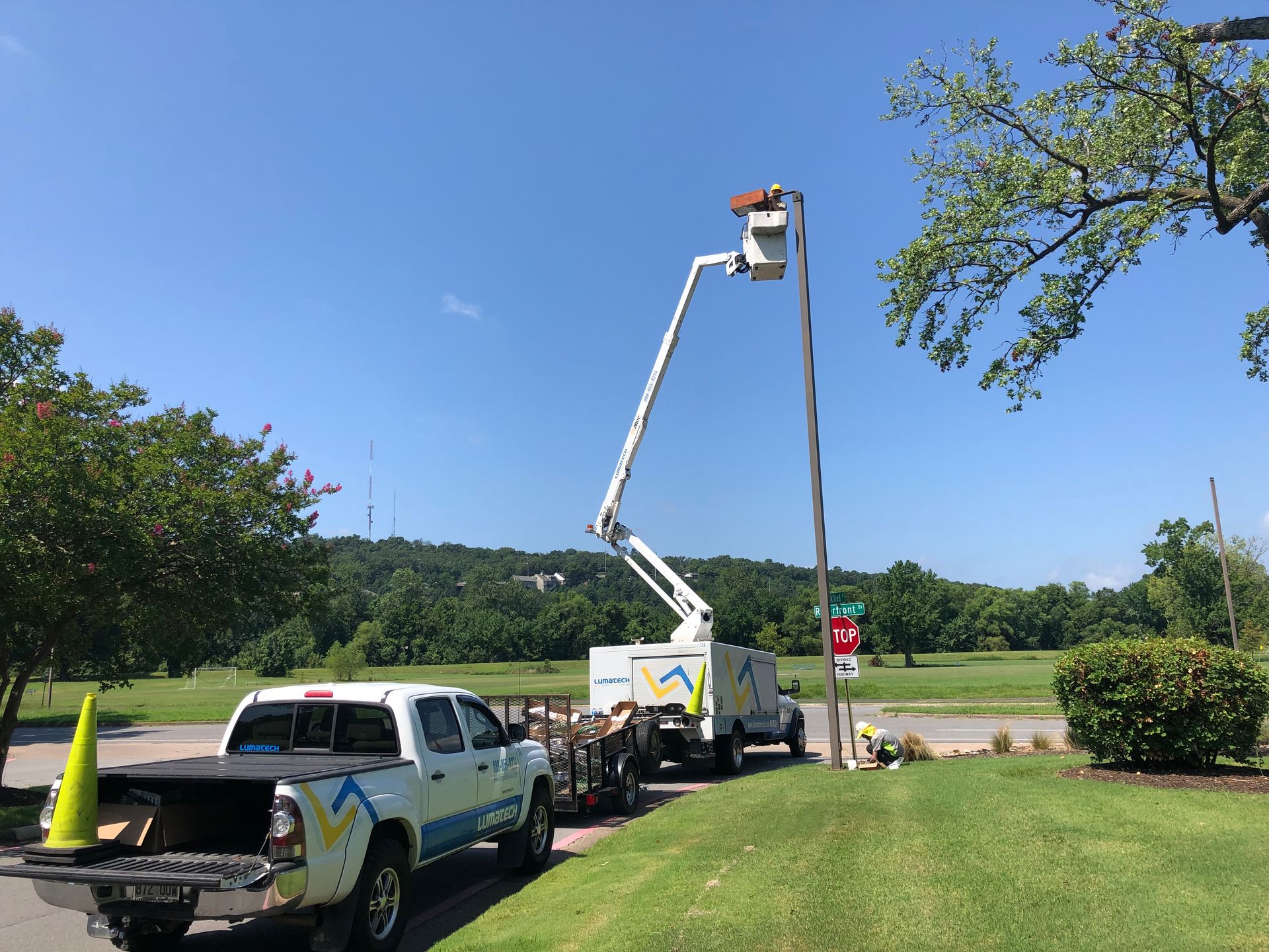 Utility worker in bucket lift trimming a tall pole beside a white truck on a sunny roadside.