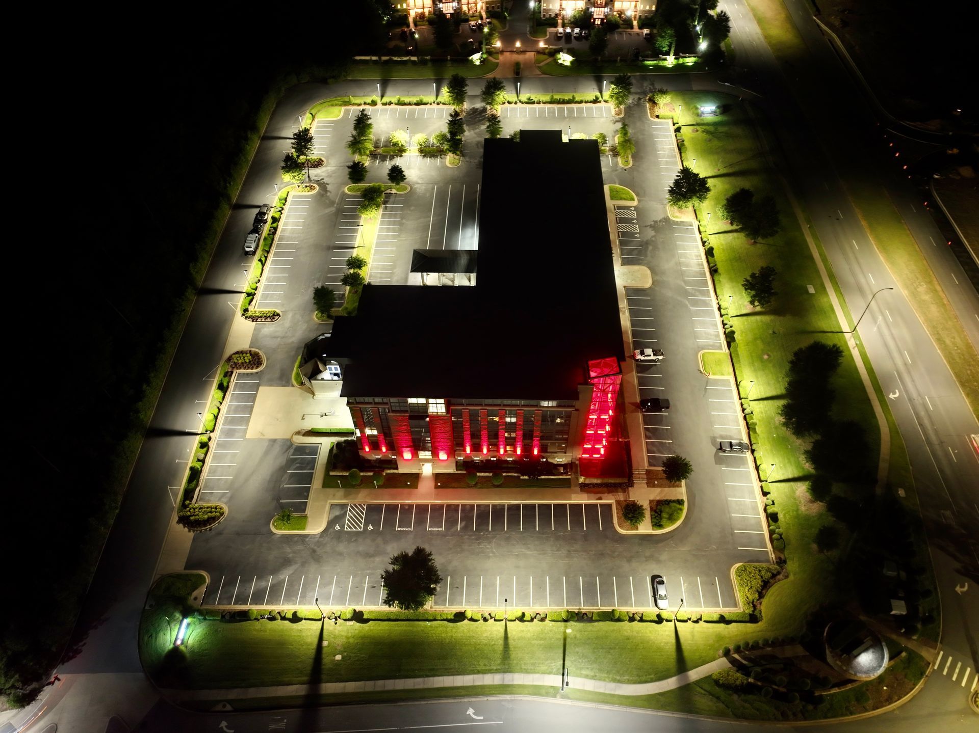 Aerial night view of a lit office building and parking lot, with red lights on the front entrance