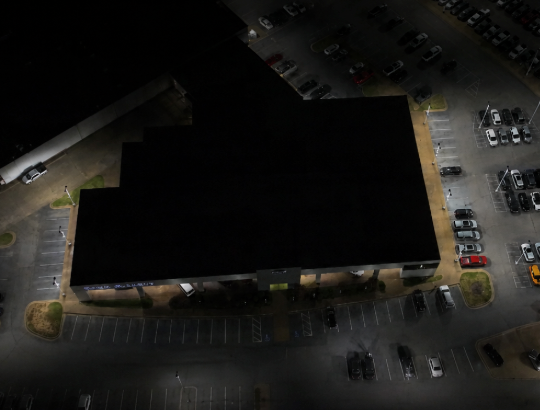 Aerial night view of a dark-roofed building surrounded by a lit parking lot with parked cars.