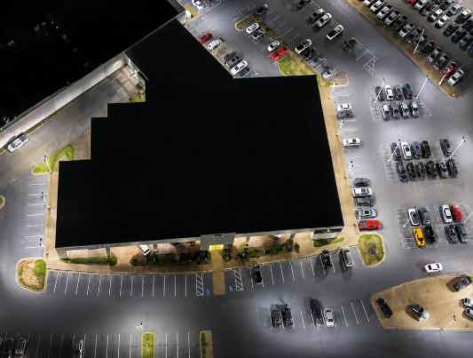 Aerial view of a large building at night surrounded by a lit parking lot with many parked cars.