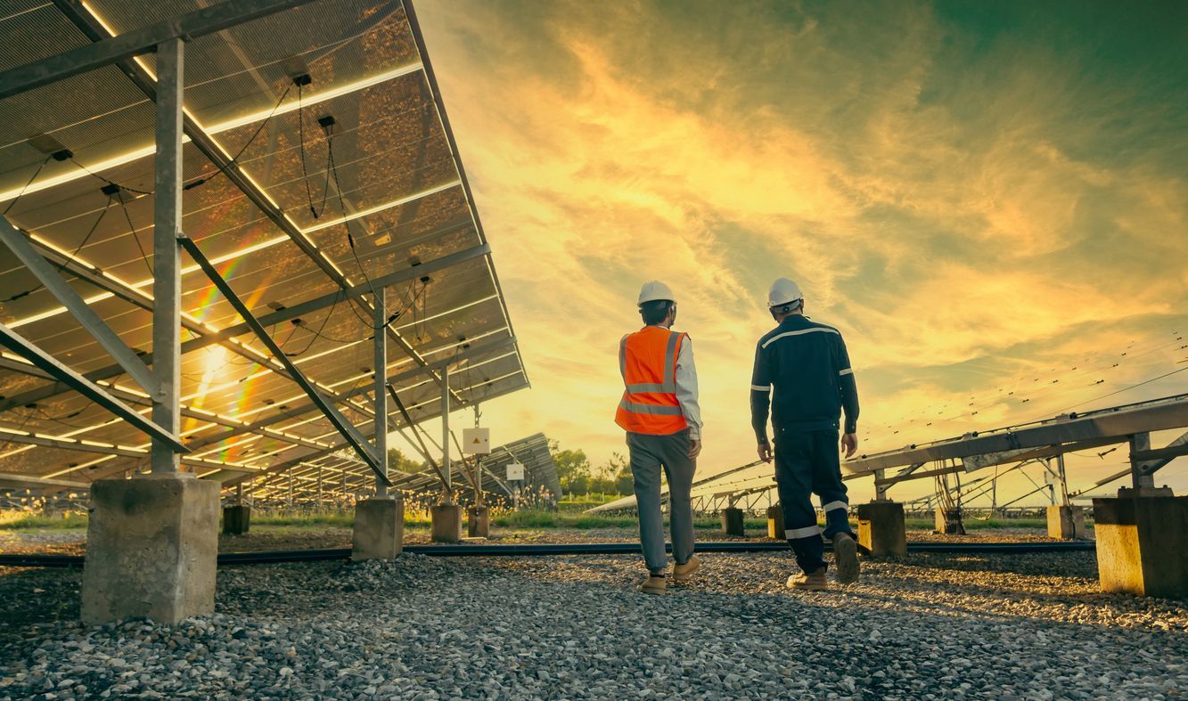 Two workers in safety gear walk beside solar panels at sunset on a gravel site.