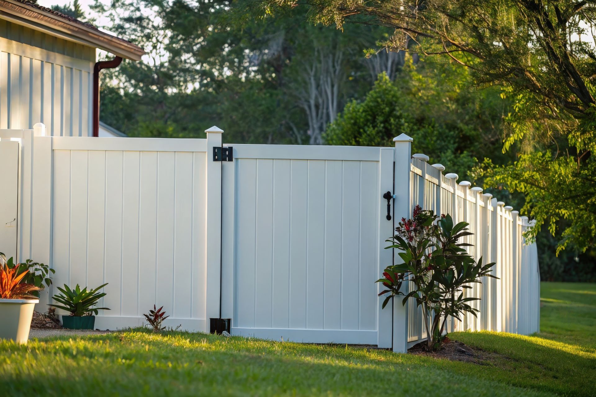 White vinyl privacy fence with a gate and plants along a landscaped yard. White vinyl privacy fence with a gate and plants along a landscaped yard.