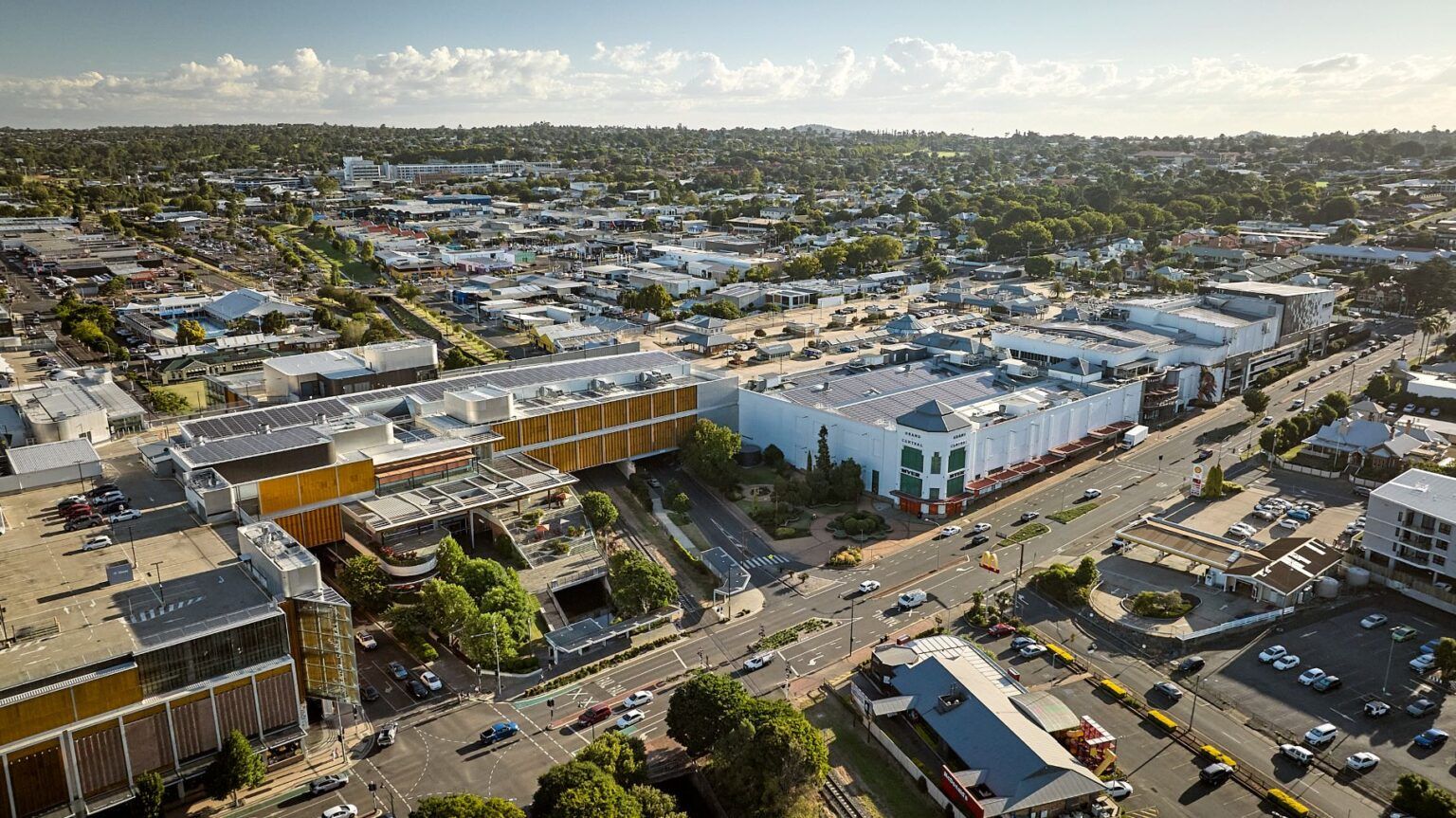 Aerial view of a city with buildings, roads, and vehicles; trees and sky in the background.