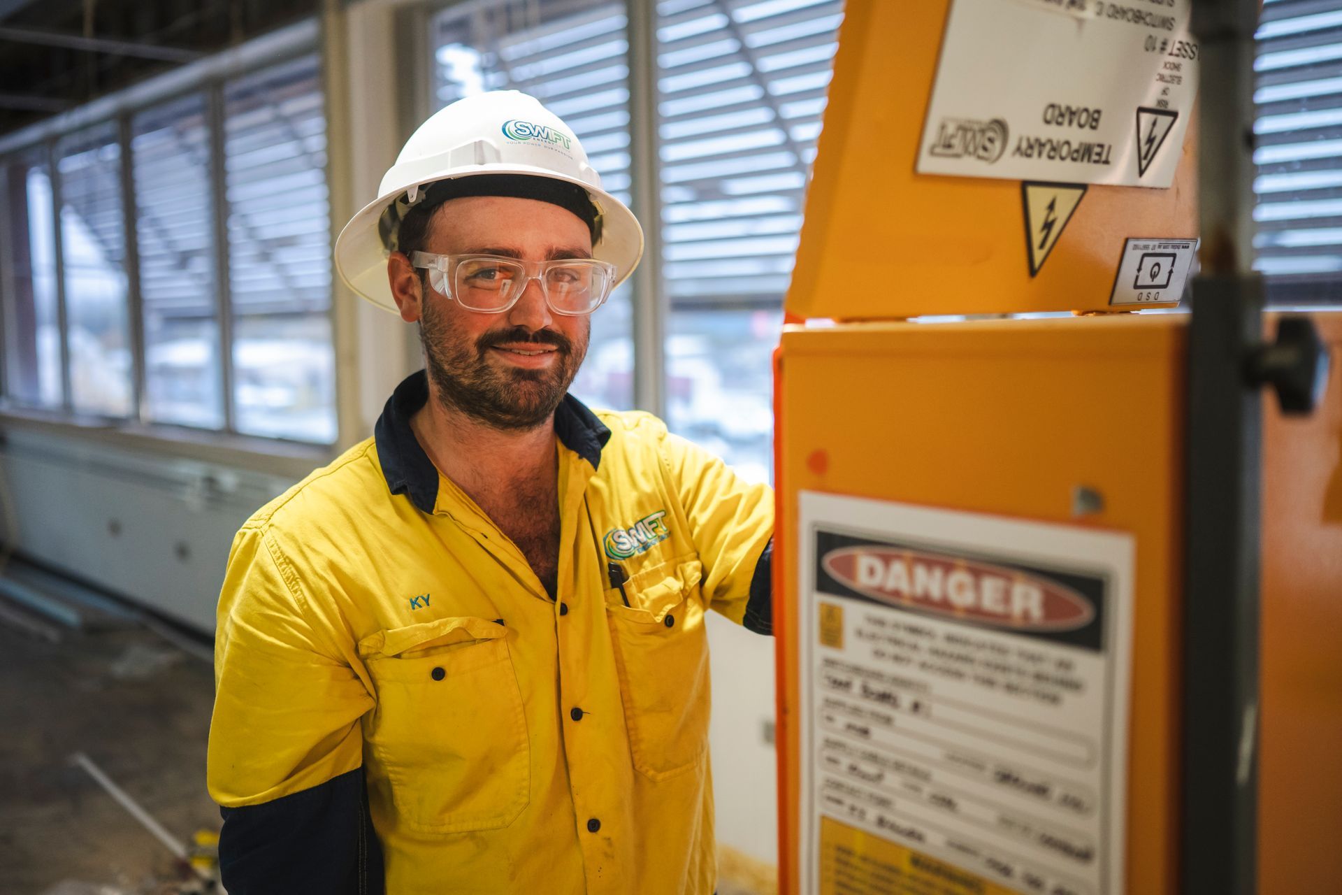 Man in yellow work clothes, safety glasses and helmet, leans against an orange machine in a workshop.