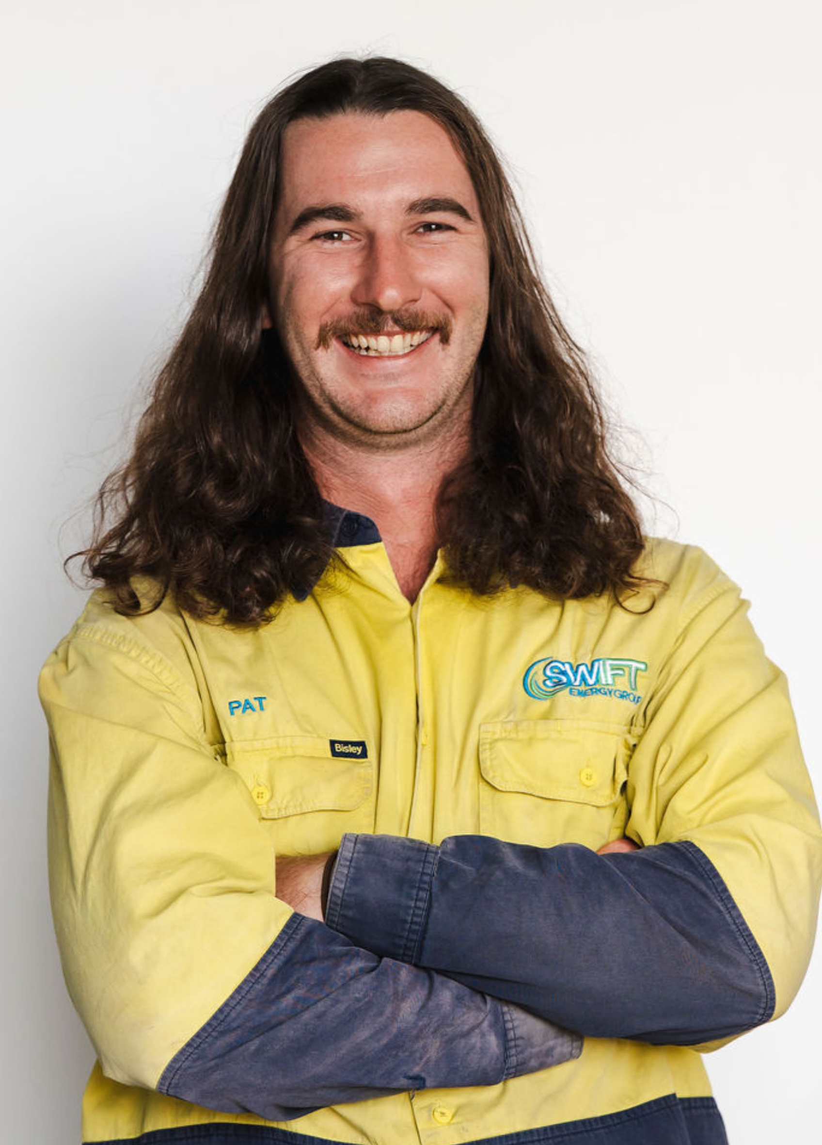 Man with long brown hair and a mustache wearing a yellow work uniform, smiling in front of a white background.