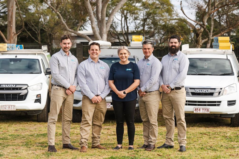 Five people posing in front of vehicles. Three men, one woman and one man. They wear work attire. Outdoors.