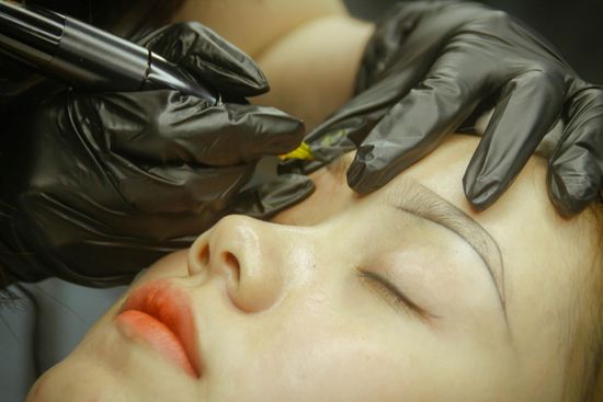 Person receiving microblading on their eyebrow by someone wearing black gloves.