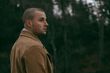 Man in a tan coat looking to the right, standing outside with a dark forest background.