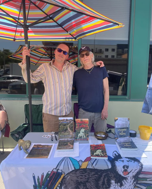 Two people stand behind a table at an outdoor craft fair under a rainbow umbrella.
