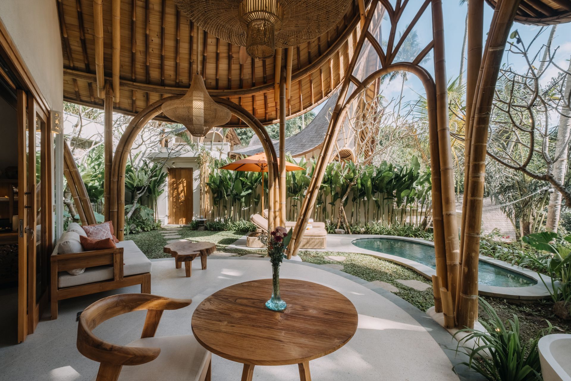 Open-air garden pavilion with round wooden table, cushioned seating, and lush plants