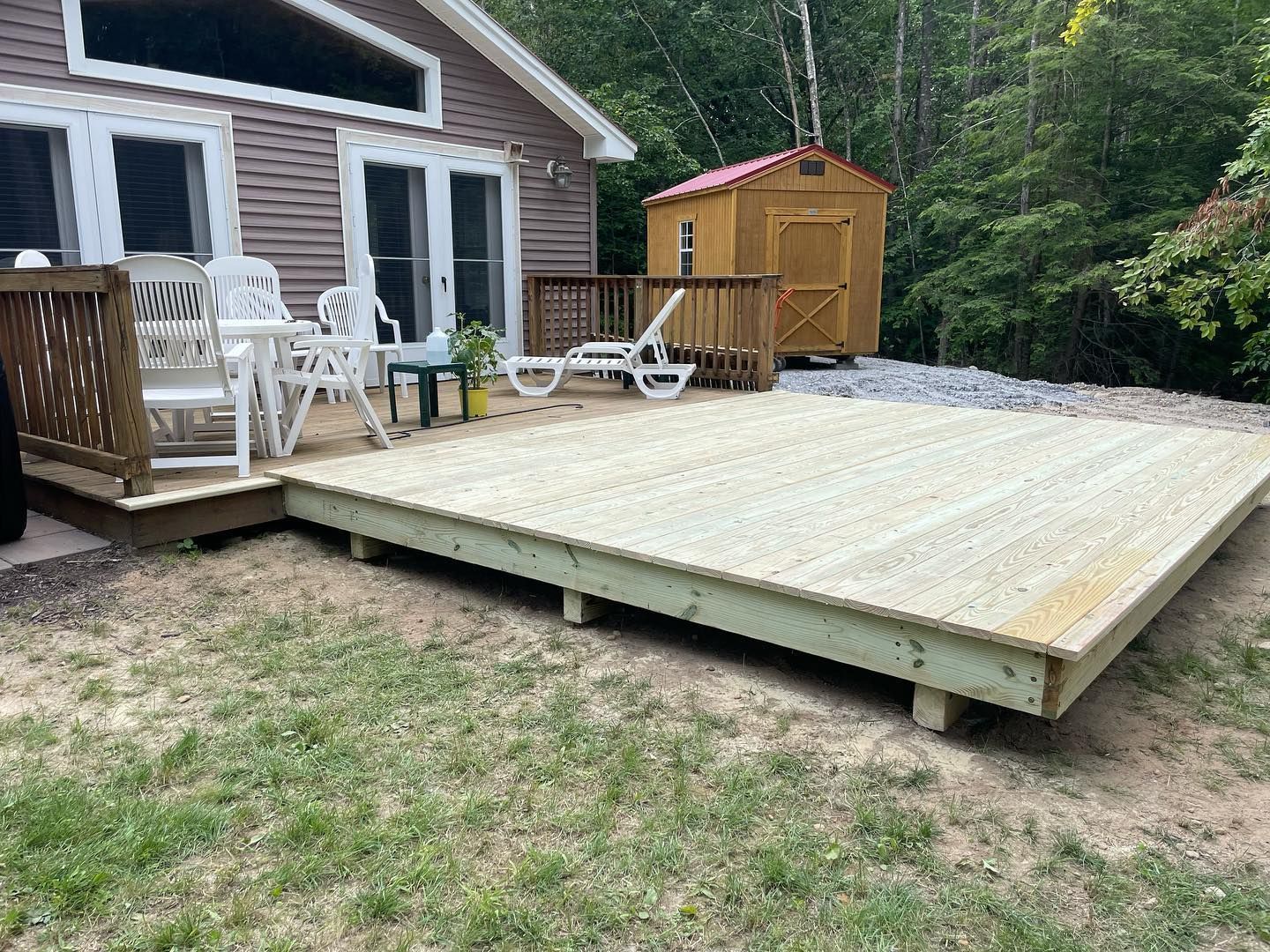 Wooden deck connected to a house with white chairs and a shed in the background. Green grass surrounds it.