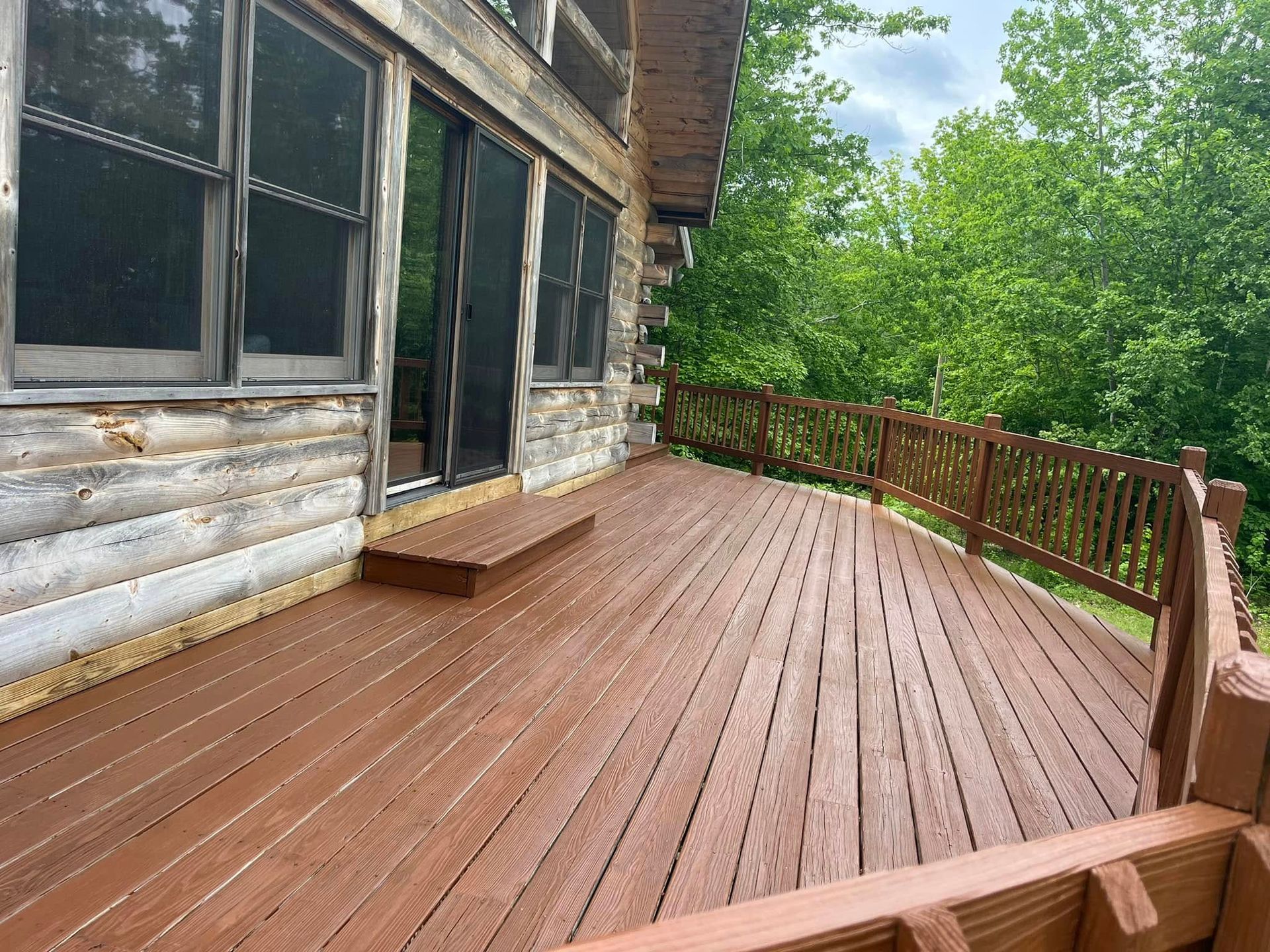 Wooden deck with brown stain and railing, attached to a rustic cabin. Green trees in the background.