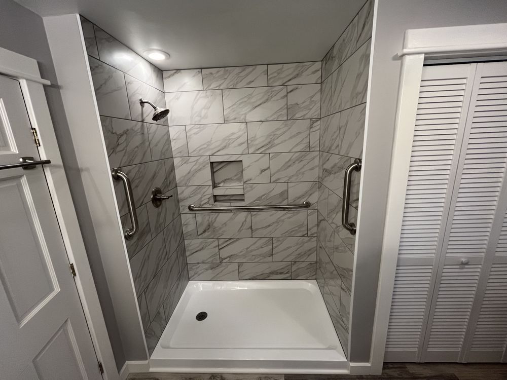 Accessible shower with marble-look tile, grab bars, and built-in niche. A white door is on the right.