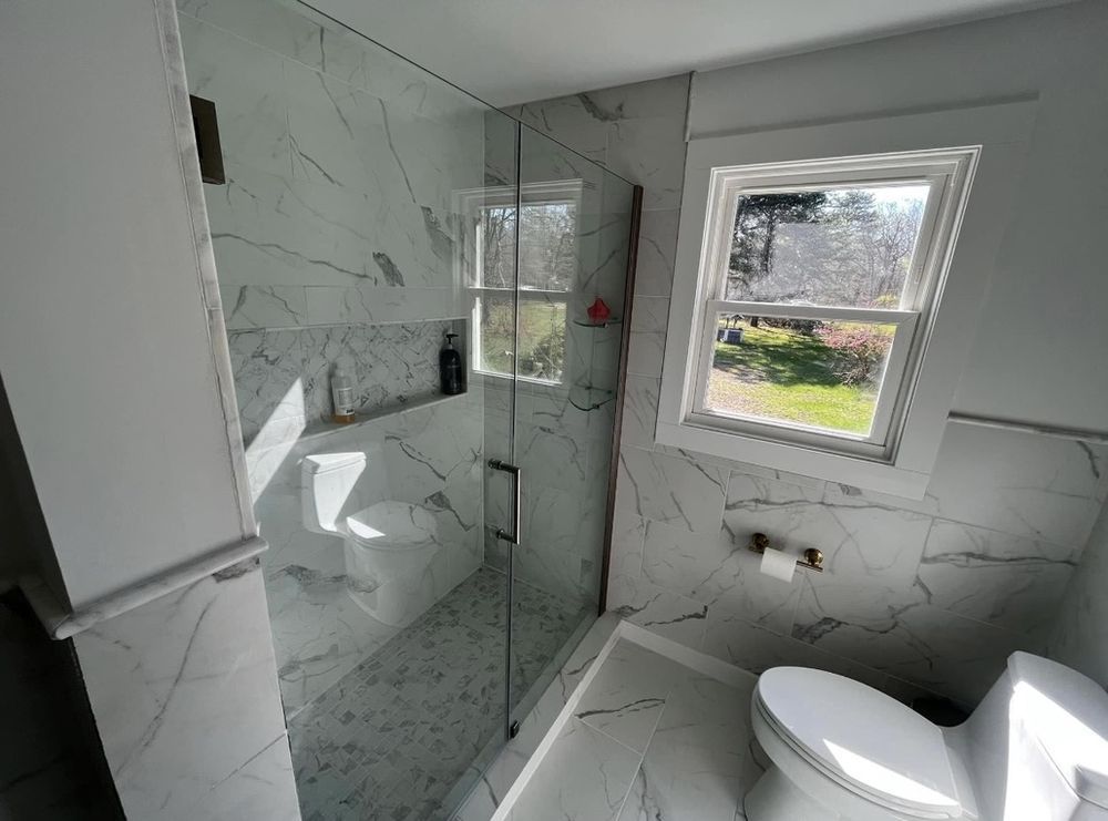 Modern bathroom with marble walls, glass shower, and toilet near a window.