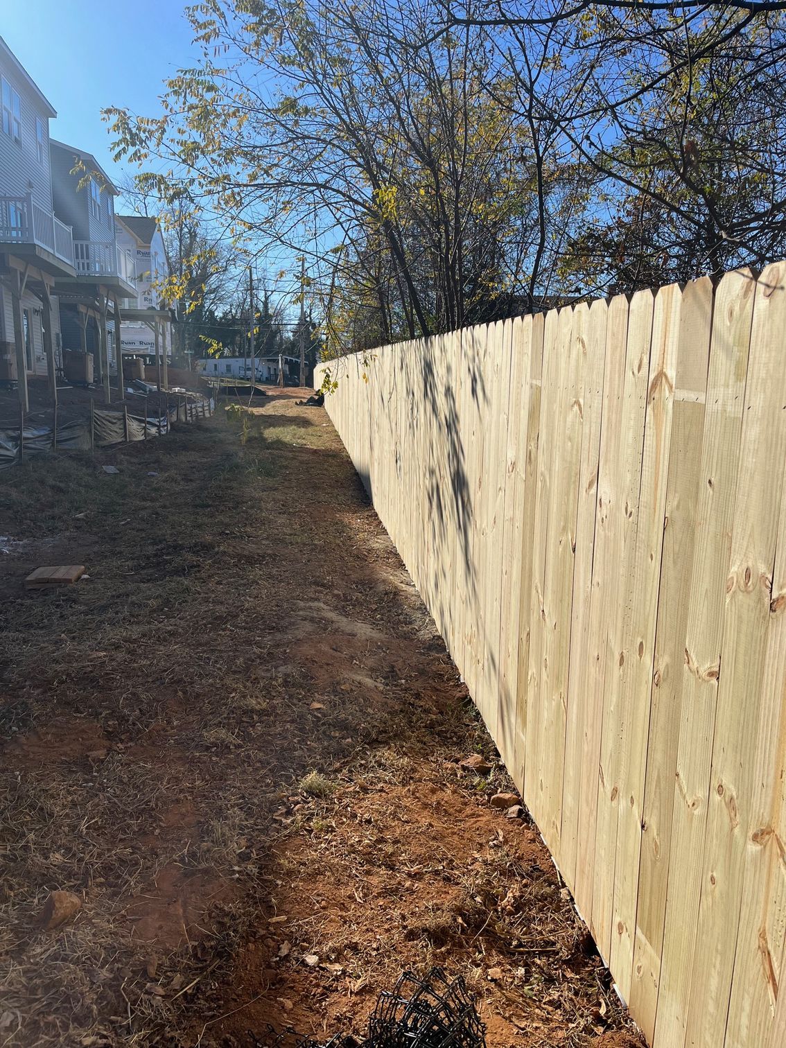 Wooden fence along a dirt path, with houses in the background and trees overhead.