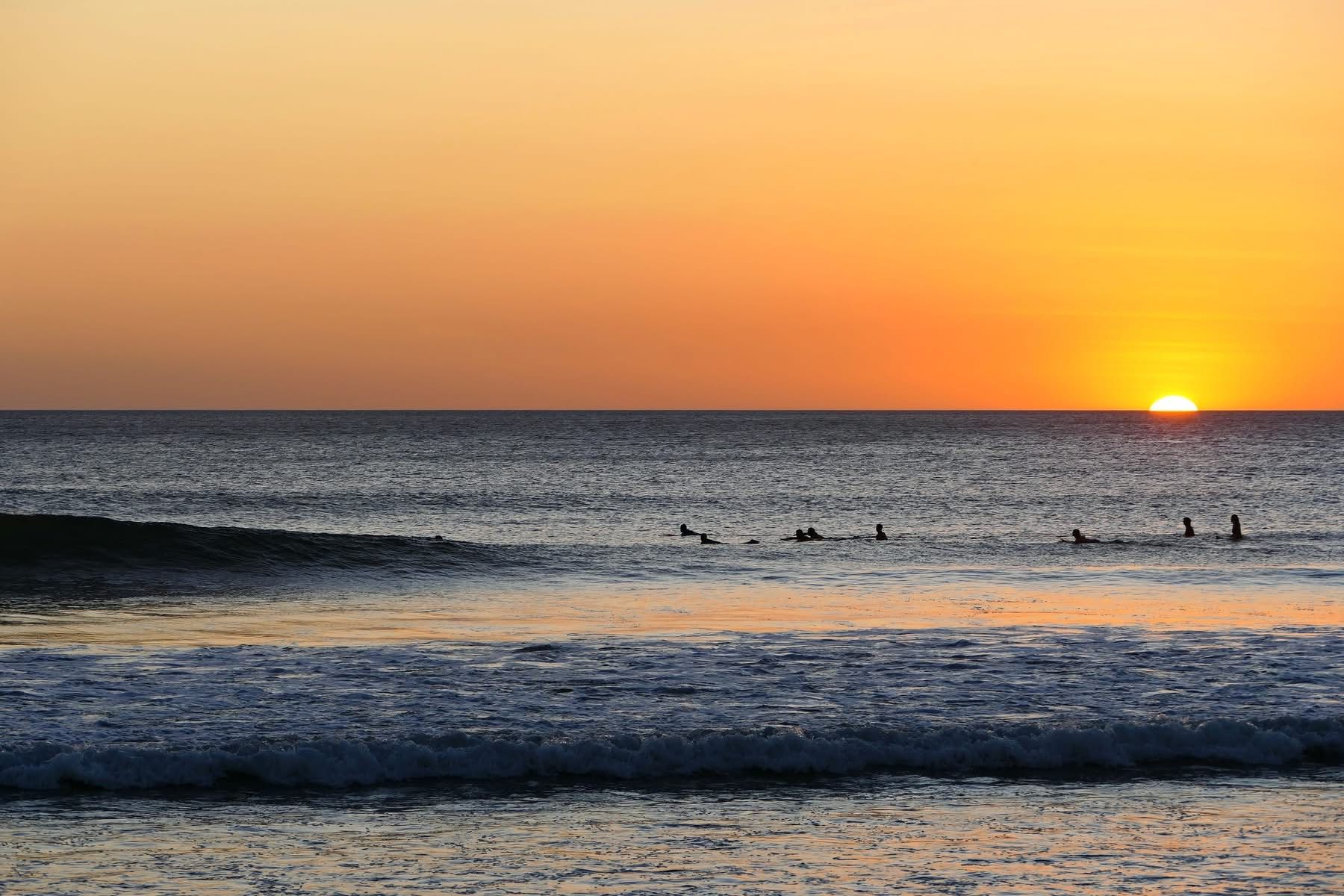 Sunset over a calm ocean with small waves and swimmers silhouetted near the horizon