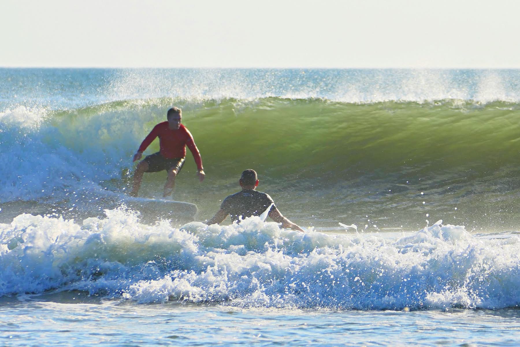 Two surfers riding a breaking green wave with white spray under a blue sky