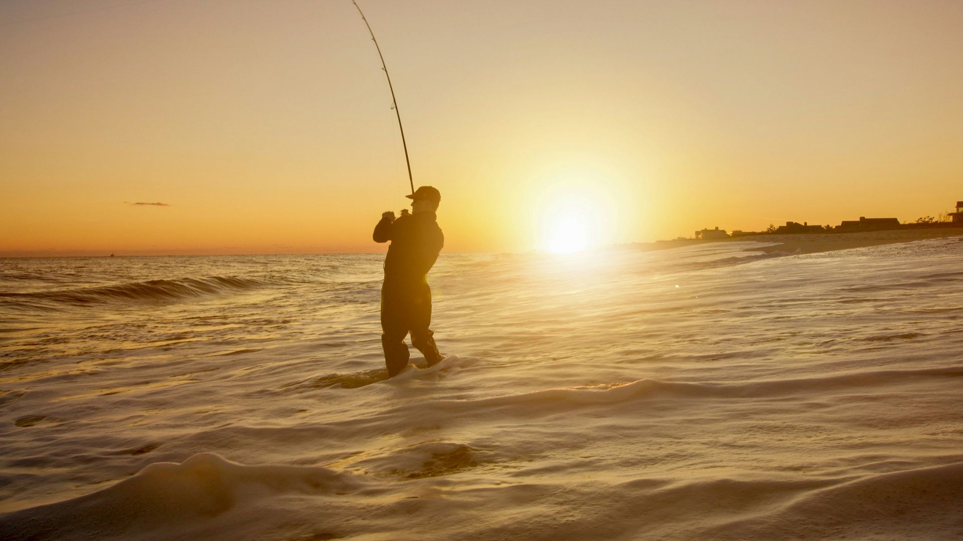 Silhouette of a person fishing at sunset on a beach, with waves and a golden sky