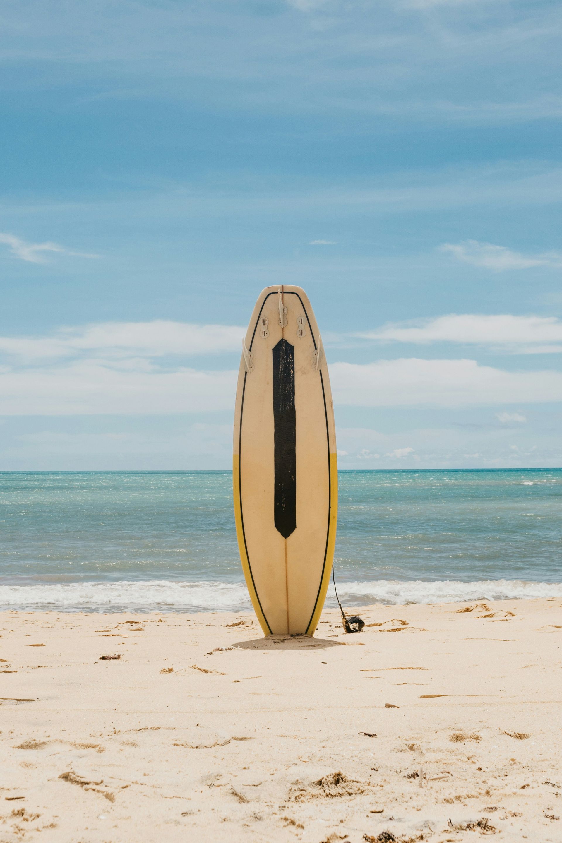 Surfboard standing upright on a sandy beach with calm blue ocean and sky behind it
