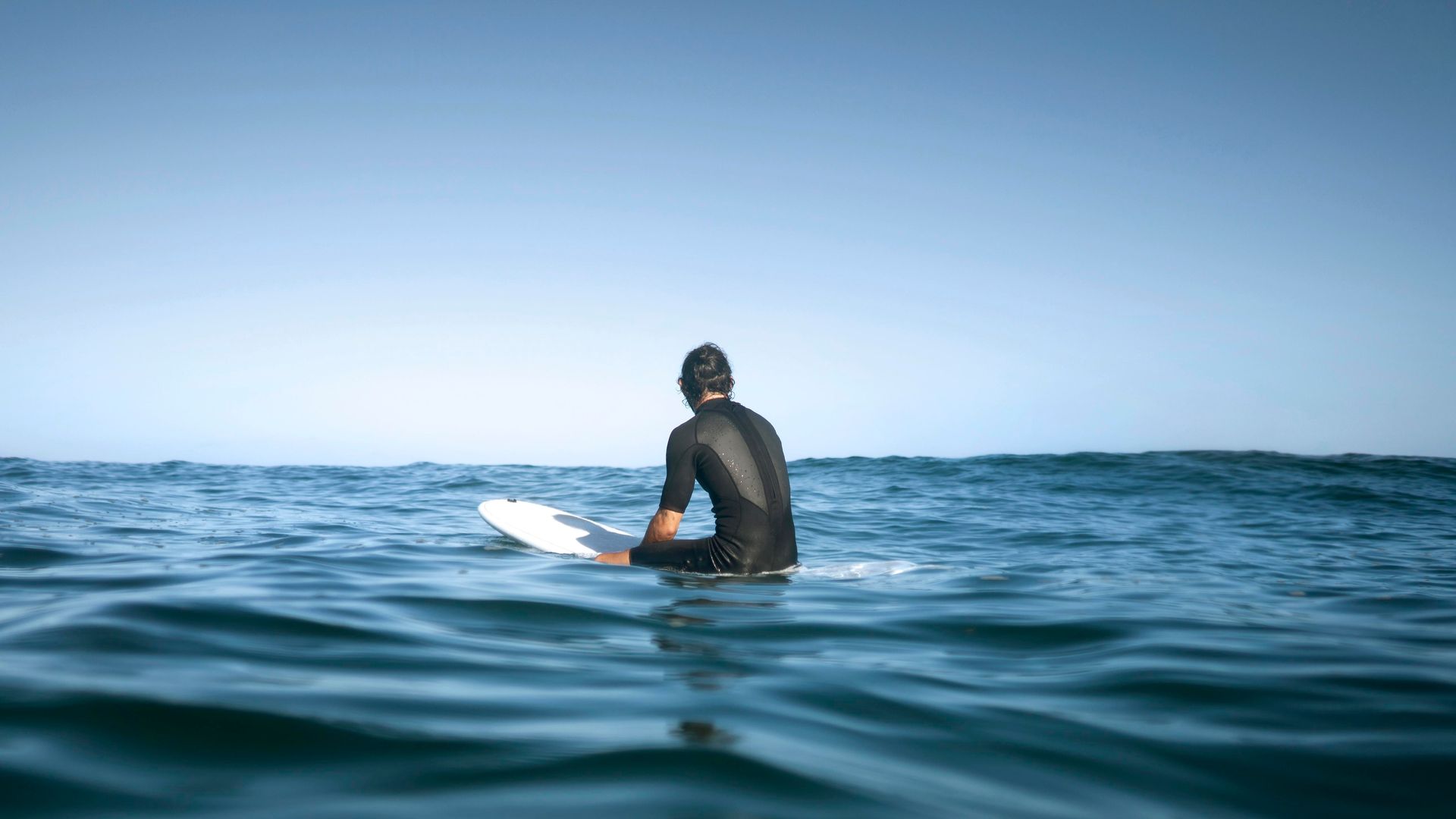 Surfer sitting on a white board in calm blue ocean water, seen from behind under a clear sky