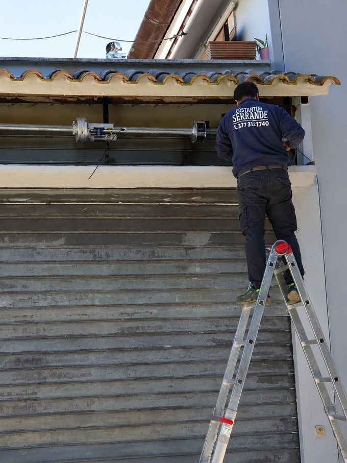 Un operaio su una scala ripara un tetto sopra un muro grigio di un cortile sotto un cielo azzurro e limpido.