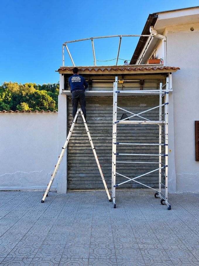 Un operaio su una scala ripara la serranda di un negozio sotto la grondaia del tetto.