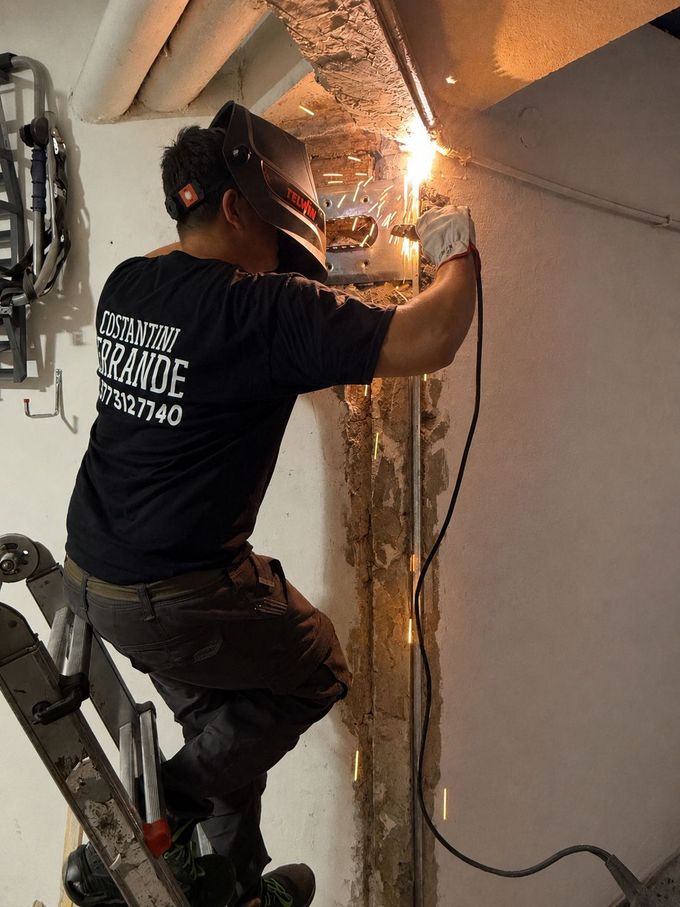 Un operaio su una scala utilizza una torcia per saldatura per riparare una struttura metallica all'interno di un edificio; le scintille volano ovunque.