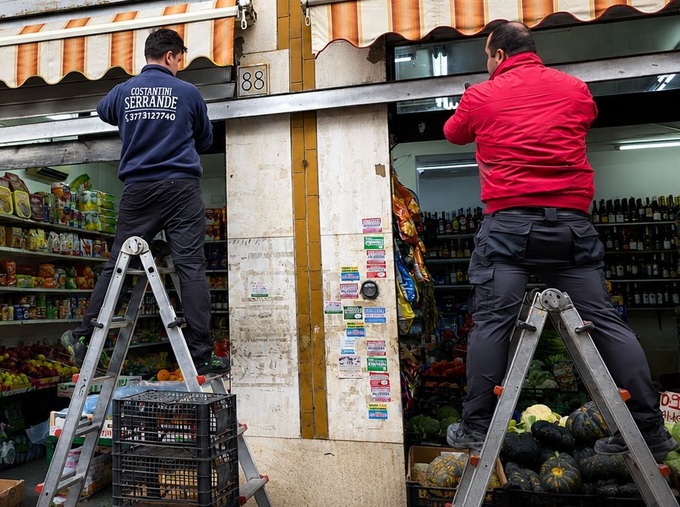 Due operai su delle scale installano una tenda da sole sopra una pila di casse di prodotti ortofrutticoli.