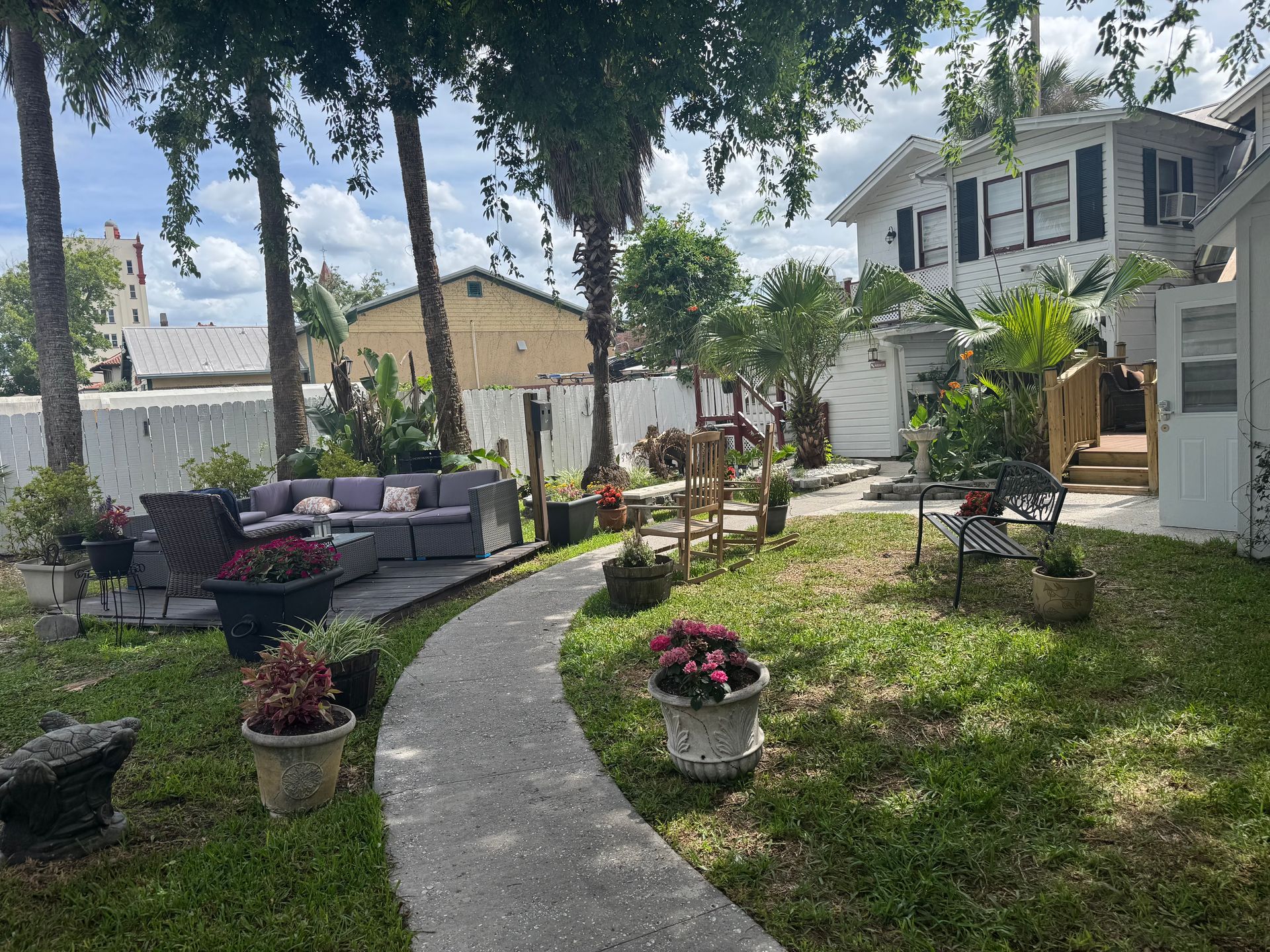 Backyard patio with curved stone path, potted flowers, seating area, and a white house under trees