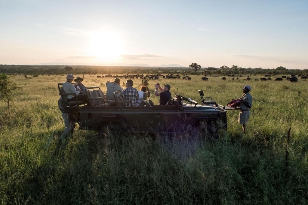 Tourists in a jeep on safari in an open field with a herd of animals in the background at sunset.