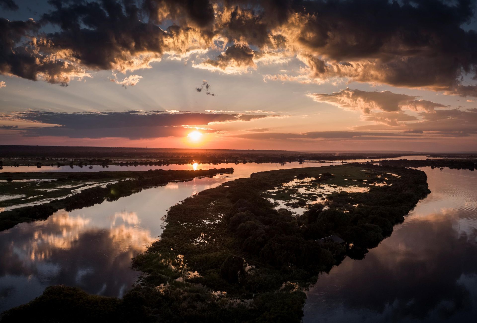 Sunset over a river with reflections of clouds and trees. Sun peeks through dramatic clouds, casting orange light.