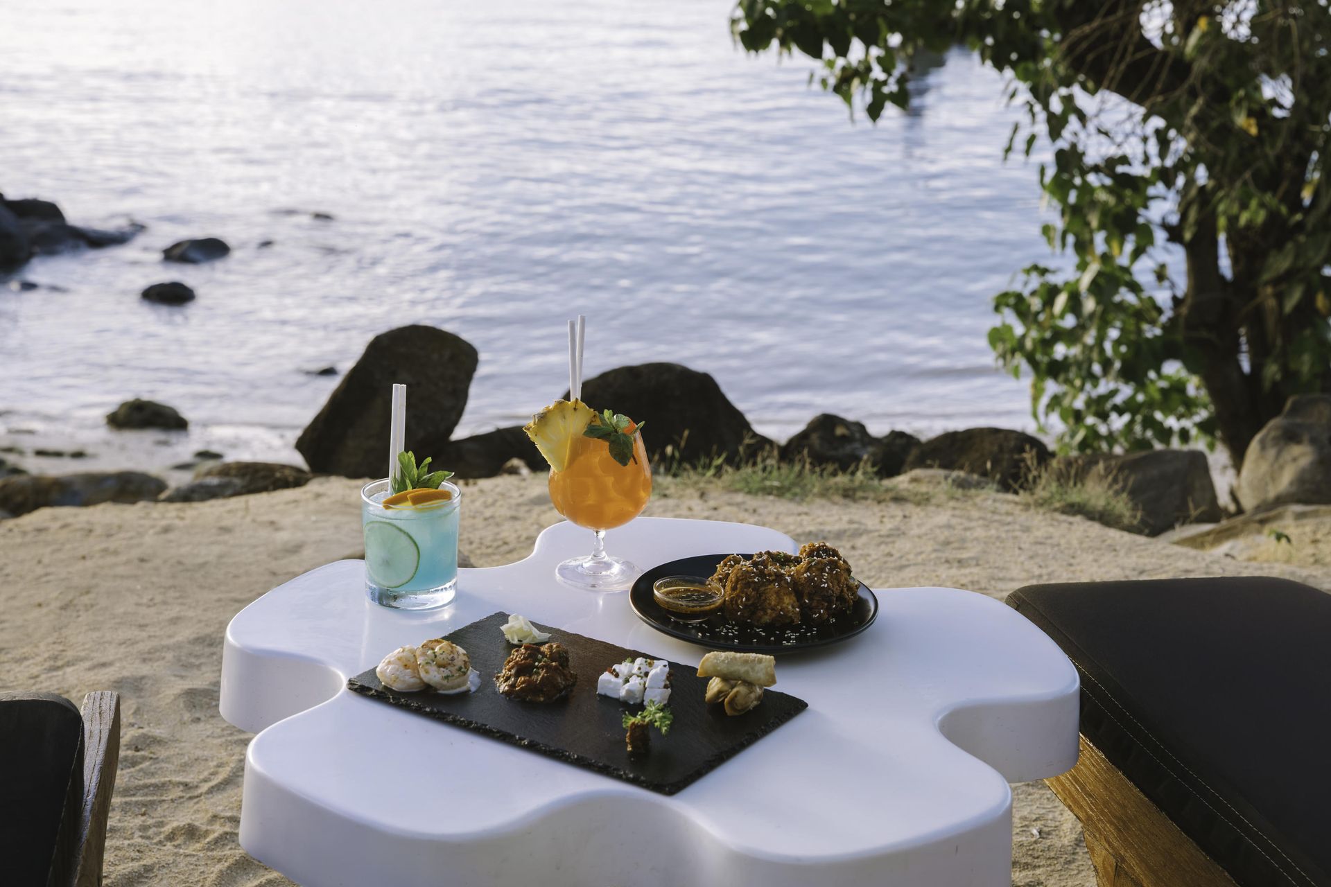 Cocktails and snacks on a white table on a beach; ocean in background.