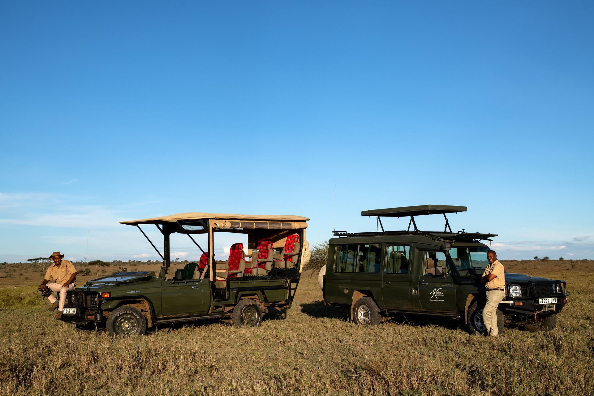 Two safari vehicles on a grassy plain, one with people seated and one with a person standing beside it under a blue sky.