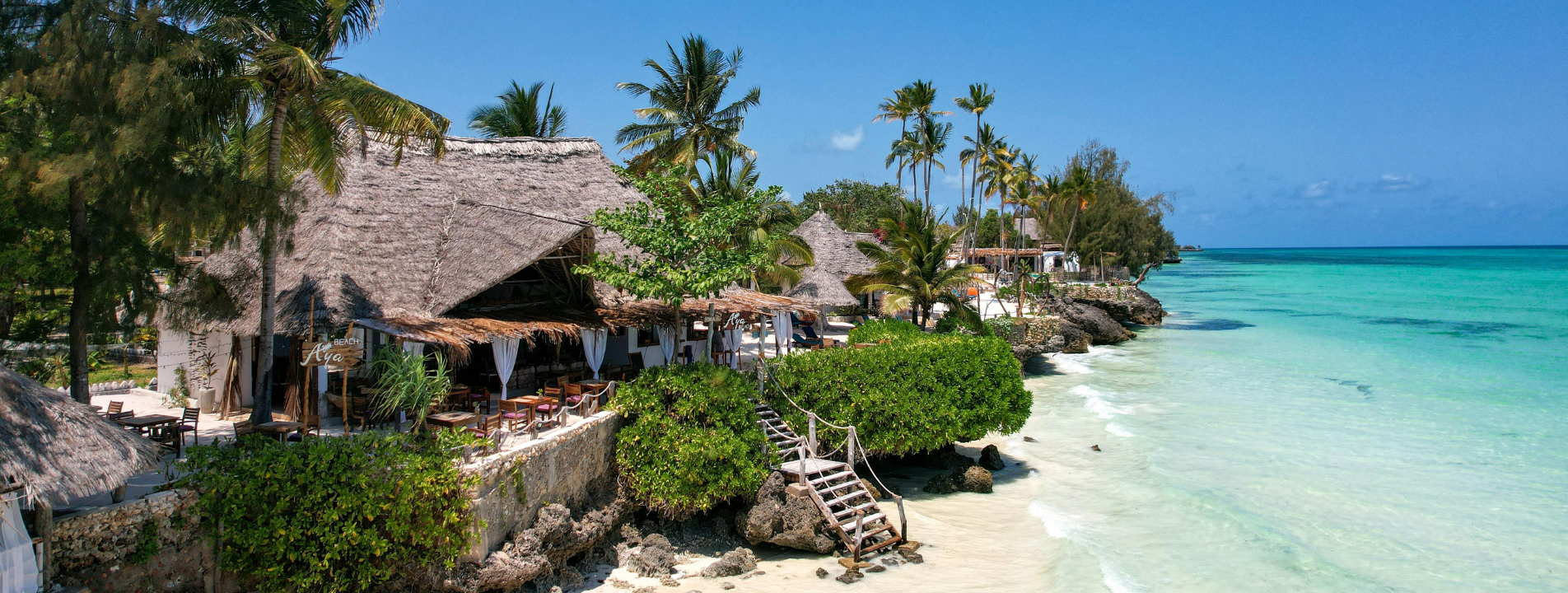 Beachfront resort with thatched roofs, turquoise water, and clear blue sky.