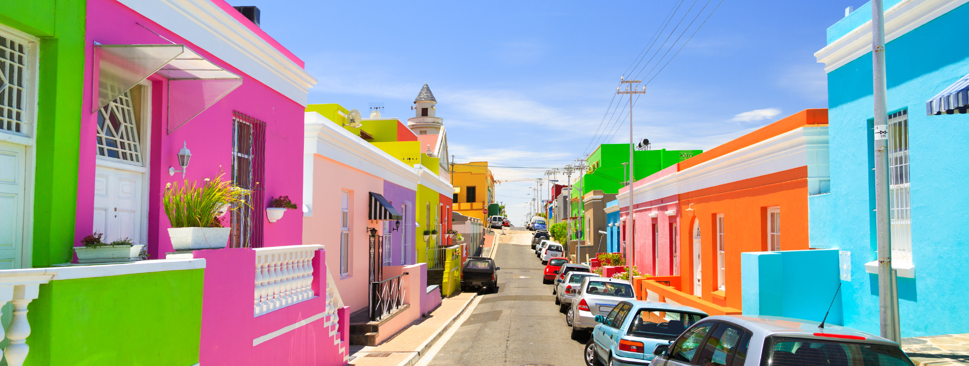 Colorful houses line a street, with parked cars. Blue sky. Bo-Kaap