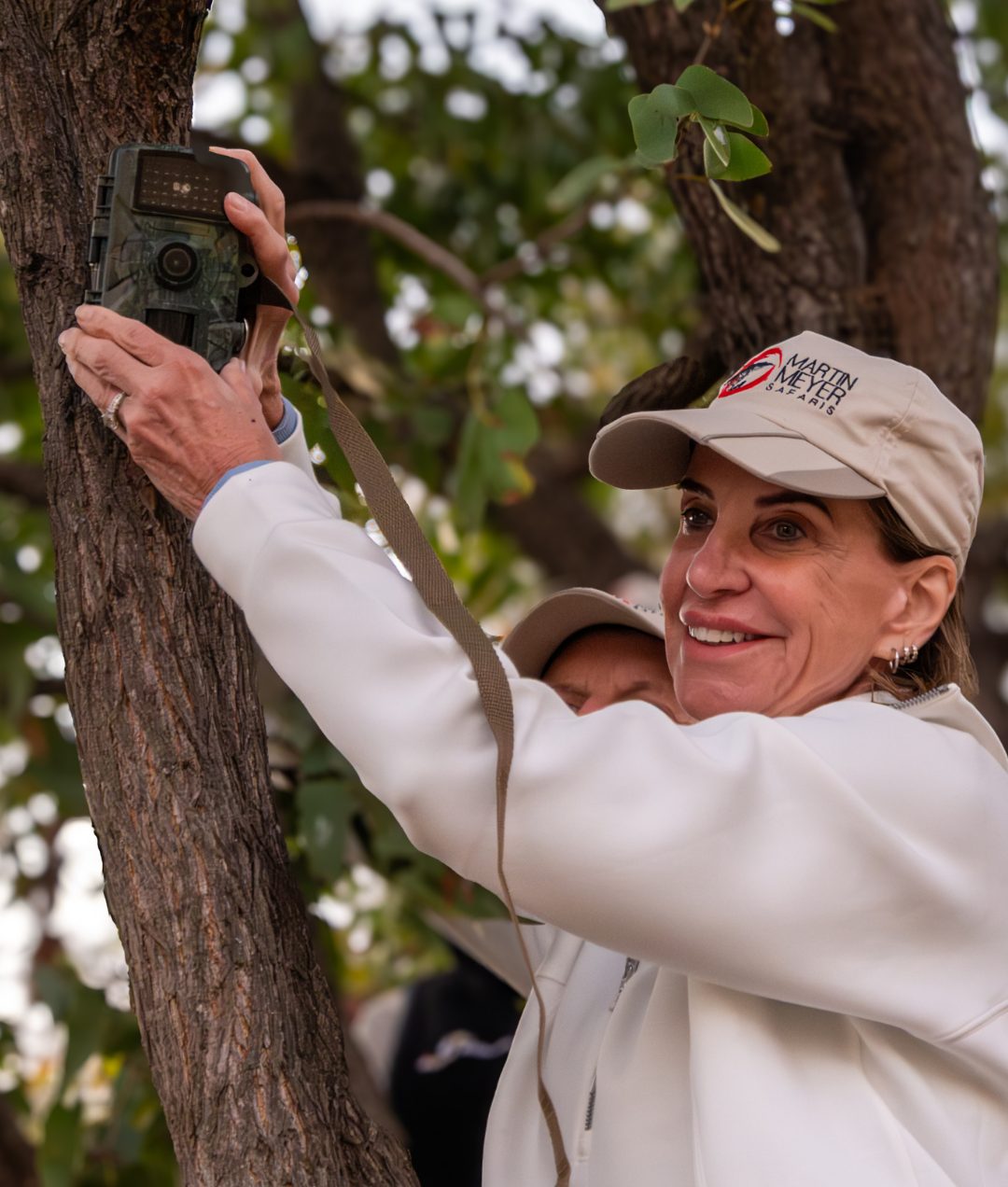 Woman in cap attaching a camera to a tree. She smiles, setting up in an outdoor setting.