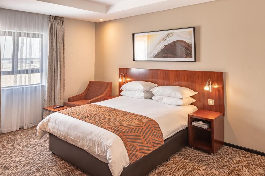Hotel room with a bed, armchair, window, and abstract artwork. Brown, white, beige color scheme.